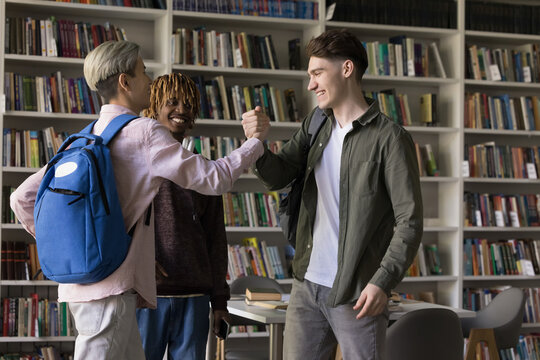 Cheerful young student guys meeting in university library for studying together, giving greeting handshakes, smiling, laughing, standing at table, bookshelves in background - Powered by Adobe