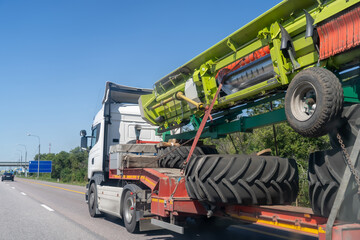 Heavy industrial truck with low side on low-frame platform transports disassembled big green combine harvester along ordinary highway on summer day.