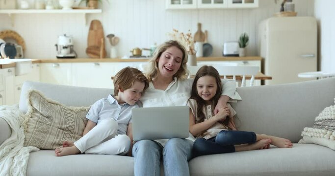 Happy blonde mother with little cute son and daughter resting on sofa with laptop, enjoy new vlog, watching on-line movie through streaming e-services, spend carefree weekend leisure use modern tech