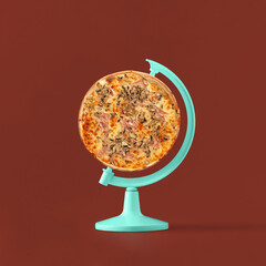 Pastel blue globe stand with whole pizza on isolated maroon colored background. Minimal abstract...