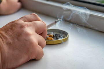A hand with a cigarette of a man smoking by the window.