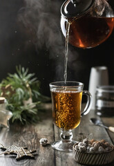 Tea is poured into a glass from a glass teapot, pouring process with steaming and Christmas decoration on wooden table. Winter time