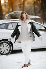 Happy caucasian woman in winter coat and mittens near car with illuminated Christmas tree on a rooftop on nature in snowy forest. Concept of celebrating New Year holidays. Idea of Xmas mood and fun