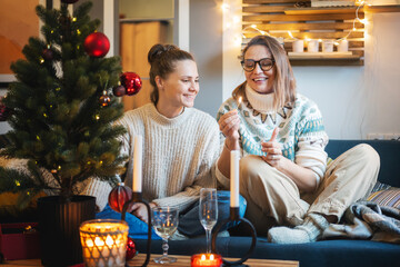 Happy lesbian couple in love in warm sweaters lighting christmas candles while sitting at home on sofa next to christmas tree