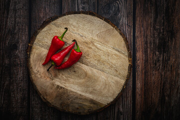 Three hot chili peppers on a round wooden cutting board on a dark wooden table.