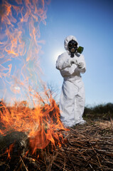 Unrecognizable person burning old dried grass in field. Ecologist holding matchbox and setting fire to dry grass under blue sky. Scientist researcher wearing protective radiation suit and gas mask.