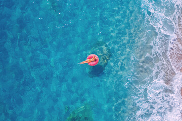 Young woman swimming on pink inflatable donut in turquoise sea with wave, aerial top view. Concept travel beach resort