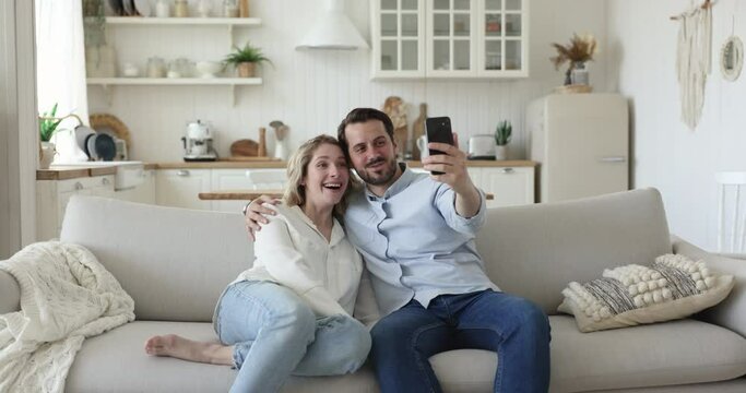 Young couple make video call using cellphone at home, enjoy virtual meeting event with family. Spouses having fun using new mobile app, making selfie pictures at dating seated on couch in living room