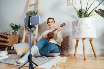 Young Woman playing ukulele guitar, recording music video on phone in modern Scandi interior home....