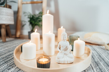Fototapeta na wymiar Wooden tray with burning candles and white Buddha statuette on the floor of modern Scandi interior. Zen Composition for meditation, yoga practice, relaxation. Balance and calm energy flow indoor.