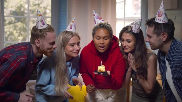 African-American woman in party hat blowing candles celebrating with noisy friends at home. Realtime