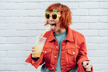 Funny Hipster fashion young woman protruding tongue in bright clothes and pineapple glasses drinking fruity tapioca bubble tea with straw on the white brick wall background. Selective focus.