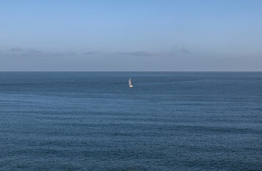Obraz premium Secluded, single white yacht sailing in the middle of of a vast dark blue ocean, some clouds and blue sky on the horizon