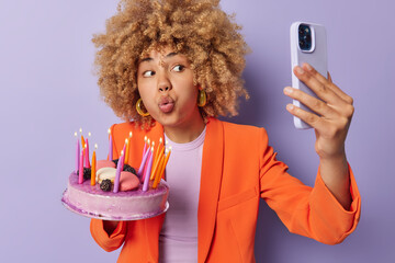 Surprised curly haired young woman poses delicious festive cake with burning candles going to make wish makes selfie via smartphone dressed in elegant orange jacket isolated over purple background.
