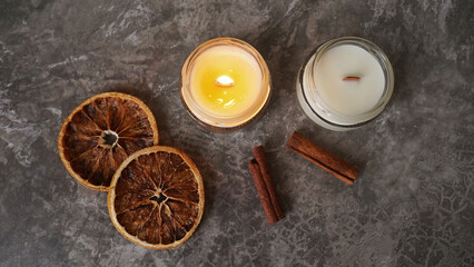 Handmade burning candle with wooden wick in white glass jar with cinnamon and dry oranges at the background, winter cozy home decoration