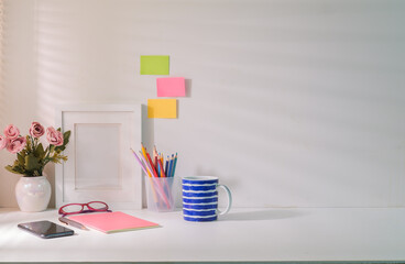 Comfortable workplace with picture frame flower pot, stationery and coffee cup on white table.