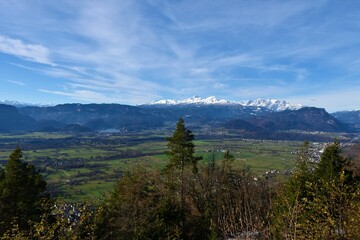 View of Triglav mountain covered in snow above forest covered hills and a plain covered in fields and Bled lake in Gorenjska, Slovenia