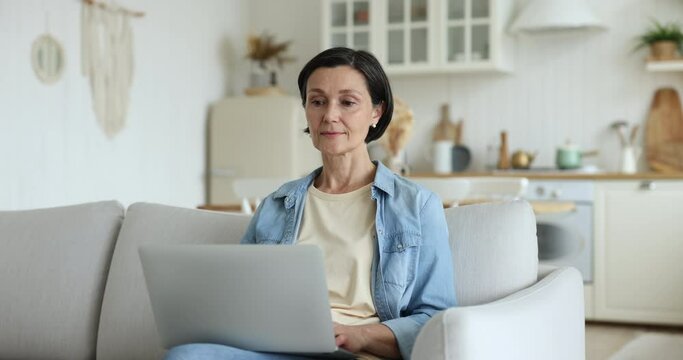 Mature woman sits on sofa with laptop get special shopping deals, big discounts, enjoy great commercial offer, celebrate on-line lottery win, bet victory feels happy. Black Friday, good internet news