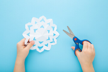 Little child hands holding scissors and cutting white snowflake shape from paper on light blue table background. Pastel color. Making decoration elements for winter festive. Closeup. Top down view.