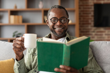 Happy mature black guy in glasses enjoys cup of coffee and reads book in free time in living room...