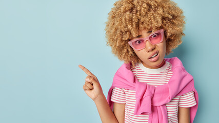 Displeased curly haired woman points index finger with unhappy expression shows something unpleasant wears pink sunglasses striped t shirt jumper over shoulders demonstrates place for your advert