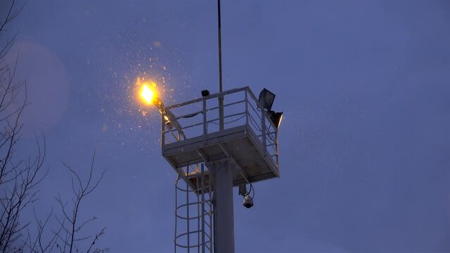 Industrial tower supporting flickering floodlight and CCTV camera at winter snowy night.