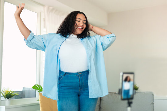 African american plus size female influencer recording video on smartphone, dancing on camera at home