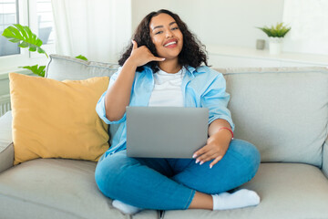 Happy black plus size woman using laptop and gesturing call me, smiling to camera, sitting on couch...