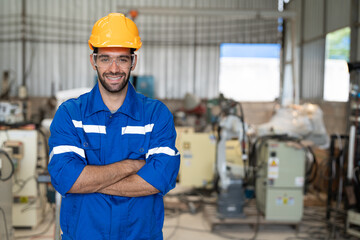 Portrait of male engineer in uniform smiling arms crossed at industrial factory. Technician man standing wearing yellow helmet safety in manufacturing workshop.