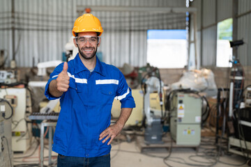 Portrait of male engineer in uniform smiling and showing thumbs up at industrial factory. Technician man standing wearing yellow helmet safety in manufacturing workshop.