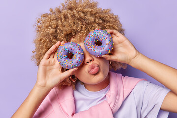 Horizontal shot of curly haired woman covers eyes with delicious glazed doughnuts keeps lips rounded poses with sweet delicious dessert poses against purple background. Unhealthy eating concept