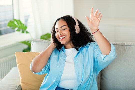 Carefree african american obese lady listening to music in wireless headphones and dancing on couch in living room