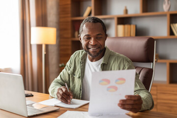 Cheerful busy middle aged black guy makes notes and works with graphs and charts, analysis...