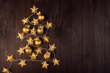 Cozy dark christmas background with golden christmas tree shape of decoration - balls, stars on glowing garland on brown wooden table, top view.