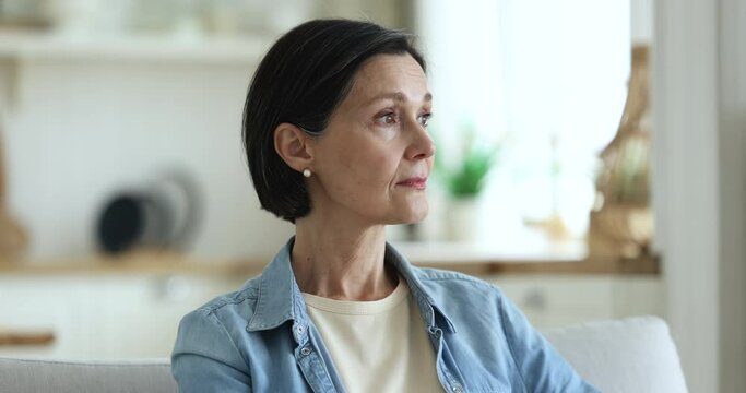 Close up head shot of attractive calm pensive middle-aged woman staring aside and thinking standing posing alone in domestic kitchen, smile looking at camera. Life on retirement, thoughts, melancholy