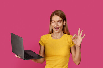 Glad adult caucasian female blonde in yellow t-shirt with open mouth winks, shows ok sign using laptop