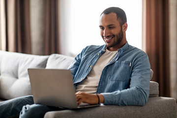 Online Correspondence. Smiling Black Man Sitting On Couch And Typing On Laptop