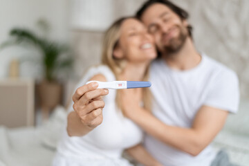 Happy caucasian spouses showing positive pregnancy test and embracing at home, selective focus. Family planning concept