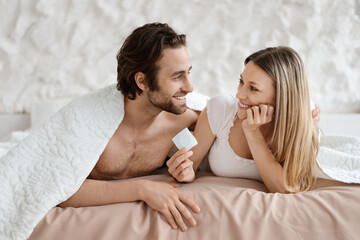 Happy loving couple lying in bed and holding condom, lovers choosing safe sex, using contraception, copy space