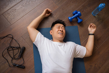 Exhausted mature korean man lying on yoga mat on floor with sports tools and bottle for water, top...