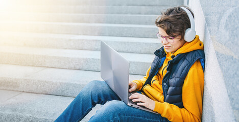 Child caucasian teen boy in yellow hoodie with headphones sitting outdoors using laptop. Smiling Programming for kids. learning on the go. Blogging surfing. Homeschooling. Video call. Copy space.