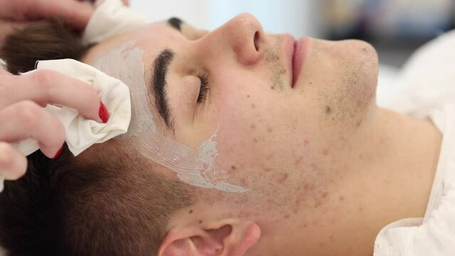 Cosmetologist washes off the clay mask with a wet napkin from man's face in spa salon. Beautician make beuty for handsome man in beauty studio. Close-up