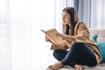 Obraz premium A young woman is reading a book while sitting on an armchair by the window.