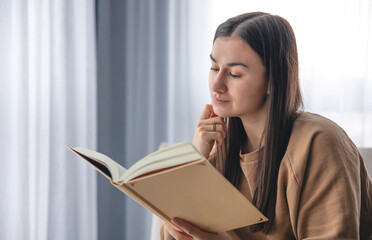 A young woman is reading a book while sitting by the window.