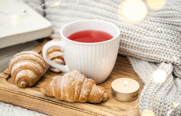 A cup of red tea, a candle, a croissant and a knitted element on a tray in bed.