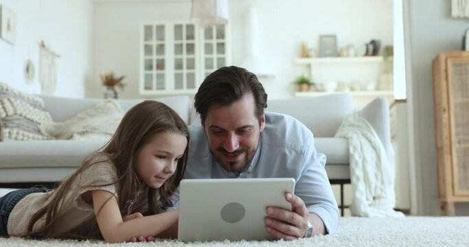 Father lying on floor with cute 6s daughter using together digital tablet at home. Happy little girl relax with loving dad, spend free time on internet watching cartoons on gadget, play on-line games
