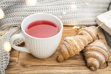 A cup of red tea, a croissant and a knitted element on a wooden tray.