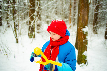 happy boy in winter bright clothes holds a snowball in his hand, walks through the snowy forest