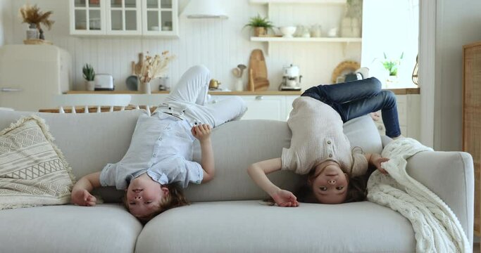 Cute little girl and boy having fun together, fooling around lying upside down on cozy sofa in living room looking carefree and happy enjoy playtime indoor. Playful children spend lazy leisure at home