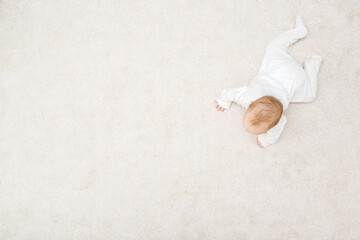 Baby in white bodysuit crawling on knee and arms on light beige home carpet background. Top down...